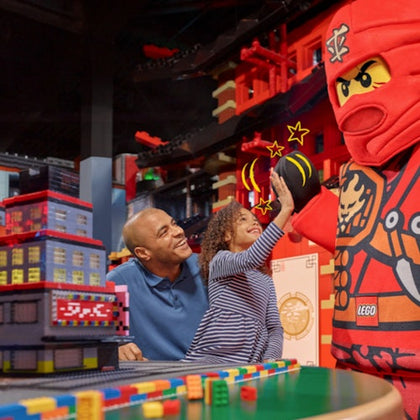 Child high-fives a person in a red LEGO ninja costume while an adult watches in a colorful LEGO-themed play area.