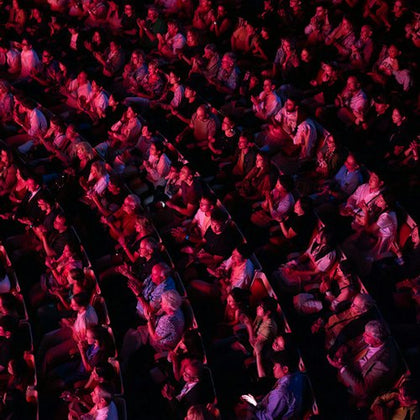 A packed theater audience seated in rows, illuminated by red lighting, facing a stage out of view.