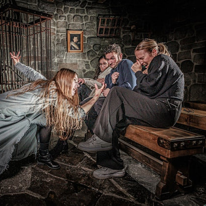 Four people in a dimly lit stone room, reacting with fear and surprise. One person in a costume is lunging towards the others.