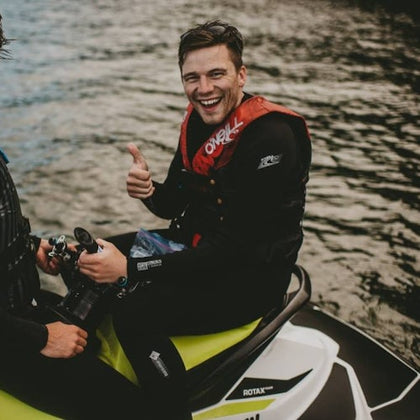 Smiling person in a wetsuit and red life vest giving a thumbs up while sitting on a jet ski on the water.