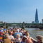 A group of people on a boat tour on a river, passing under a bridge, with a tall skyscraper visible in the background.