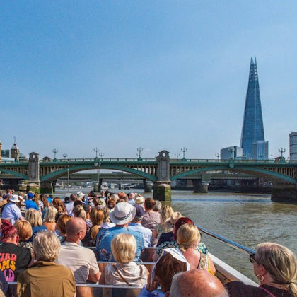 A group of people on a boat tour on a river, passing under a bridge, with a tall skyscraper visible in the background.