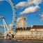 A large Ferris wheel near a historic building and a high-rise, with a river in the foreground and a partly cloudy sky.