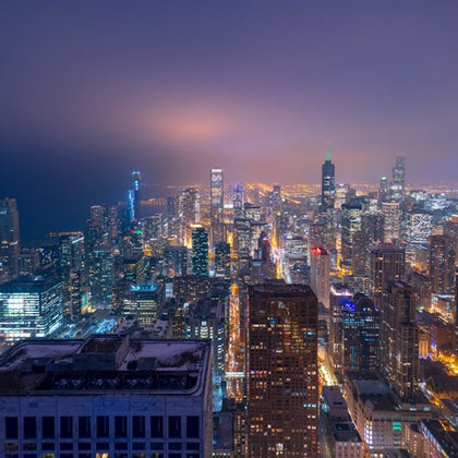 Aerial view of a city skyline at night with illuminated skyscrapers and buildings under a hazy purple sky.