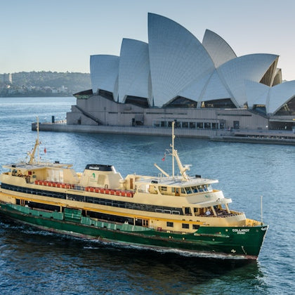 A ferry sails in front of the Sydney Opera House on a sunny day, with the shoreline visible in the background.