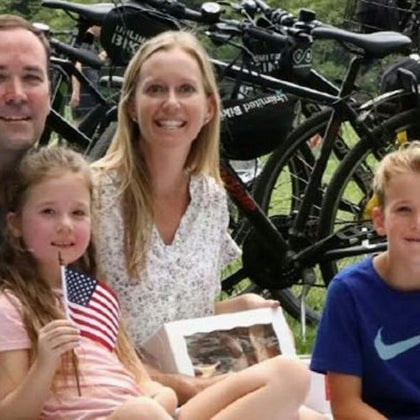 A group of people sitting outdoors near bicycles, with a young girl holding an American flag and others smiling at the camera.