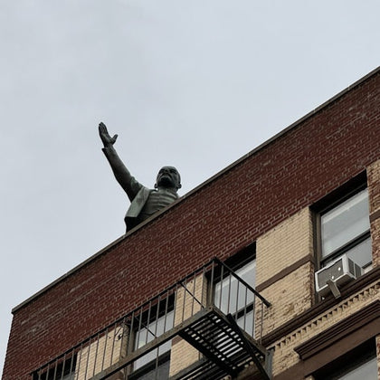 A statue of a person extending a raised arm on the rooftop of a brick building with visible fire escape and windows.