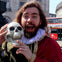 A person dressed in Shakespearean attire holding a skull, with a red double-decker bus in the background.