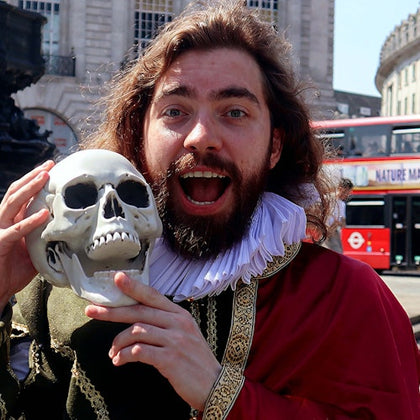 A person dressed in Shakespearean attire holding a skull, with a red double-decker bus in the background.