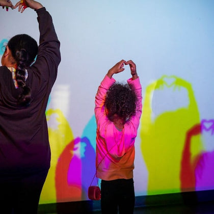Two people stand facing a wall, casting colorful shadow shapes with their raised hands illuminated by lights.