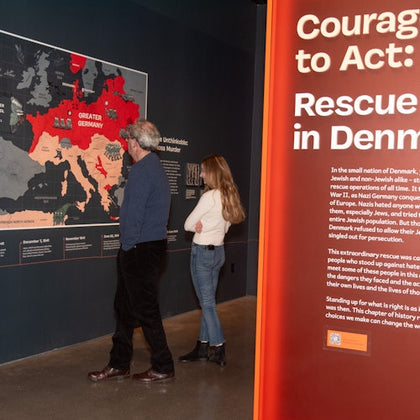 Two people view an exhibit featuring a map and text about Denmark's rescue efforts during WWII, with a large red panel displaying the title "Courage to Act."