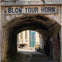 A narrow brick archway with a sign reading 'Blow Your Horn' leads to a courtyard with a green door and outdoor items.