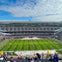 Football stadium with players warming up on the field and spectators in the stands under a partly cloudy sky.