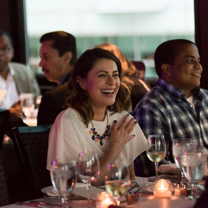 A group of people seated at a dinner table, a woman in the foreground smiling and touching her chest, surrounded by wine glasses.