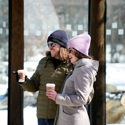 Two people in winter clothing, each holding a coffee cup, smile and stand outdoors with snow and trees in the background.