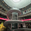 Interior of a modern public space with a spiral walkway, glass dome ceiling, and illuminated signs along the perimeter.