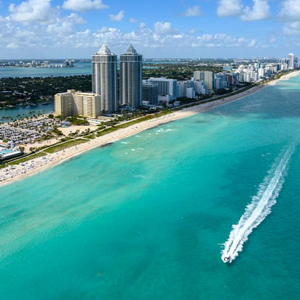 Aerial view of a coastal city with high-rise buildings, a crowded beach, and a speedboat on turquoise water under a blue sky.