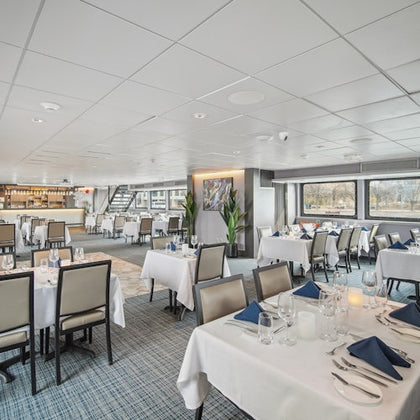 A spacious, modern restaurant with neatly set tables, white tablecloths, blue napkins, and large windows overlooking a scenic view.