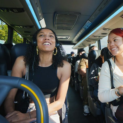 Two women smiling and wearing headphones while seated on a bus, with other passengers visible in the background.
