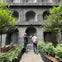 Two people standing in front of a textured wall with arched recesses, surrounded by greenery and trees.