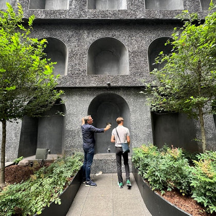 Two people standing in front of a textured wall with arched recesses, surrounded by greenery and trees.