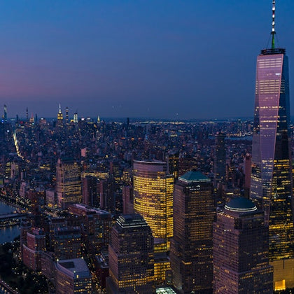 Aerial view of a cityscape at dusk with illuminated skyscrapers and a prominent tall building on the right.
