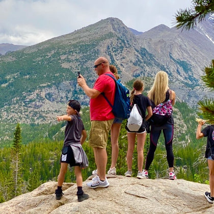 A group of people, including adults and children, stand on a rock, overlooking a mountainous forested landscape.
