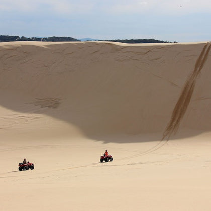 Two people riding ATVs on expansive sand dunes under a clear sky, with tire tracks visible on the slope.