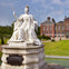 White statue of a crowned and robed figure holding a scepter, situated in front of a historic brick building with green lawn.