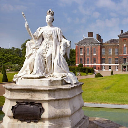 White statue of a crowned and robed figure holding a scepter, situated in front of a historic brick building with green lawn.