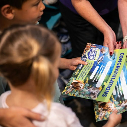 Two children receive a colorful certificate from an adult at an event, with the word 'CONGRATULATIONS' visible.