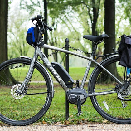 A silver electric bicycle with black accessories is locked to a post in a park with grass and trees in the background.