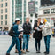 Four people stand in discussion in a busy urban plaza, holding and looking at papers. Buildings and a digital billboard are in the background.