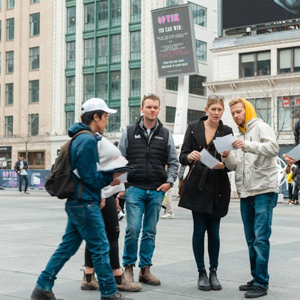 Four people stand in discussion in a busy urban plaza, holding and looking at papers. Buildings and a digital billboard are in the background.