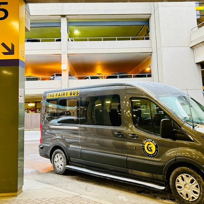 A gray van labeled "The Fairy Bus" is parked near a multi-story parking garage, with signs and lighting visible in the background.