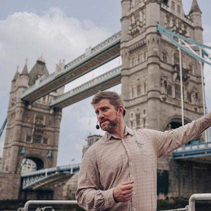 A man wearing a headset gestures while standing in front of the Tower Bridge in London on a cloudy day.