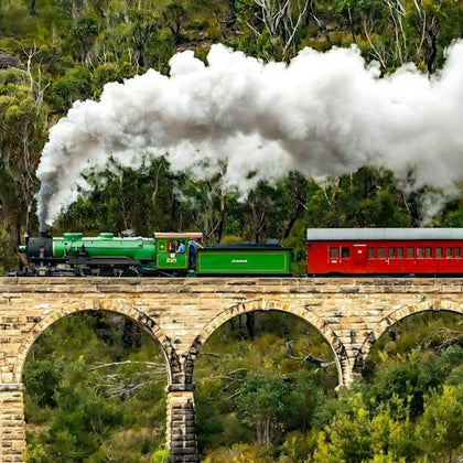 A green steam train with red passenger cars crossing a stone bridge in a forested area, emitting white smoke.