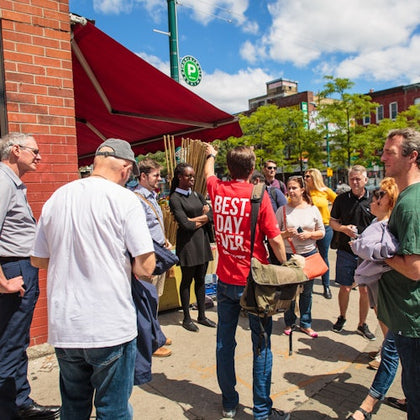 A group of people gather on a sunny sidewalk outside a red awning next to a brick building, with one person wearing a "Best. Day. Ever." shirt.