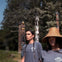 Two individuals wearing traditional attire stand outdoors near tall totem poles, with trees visible in the background.