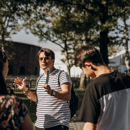 A man in sunglasses is talking to two people outdoors, holding a blue object, with trees and a building in the background.