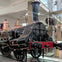 A historic steam locomotive displayed indoors, featuring black and red colors with copper details, in a museum setting.