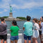People on a boat observe and photograph the Statue of Liberty on a sunny day, with the statue and skyline in the background.