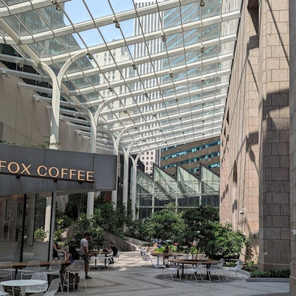 An outdoor seating area with tables and chairs under a glass roof, next to a cafe with a sign partially reading 'FOX COFFEE.'