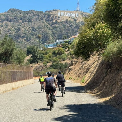 Three cyclists ride on a paved road with the Hollywood sign visible on a shrub-covered hill in the background.