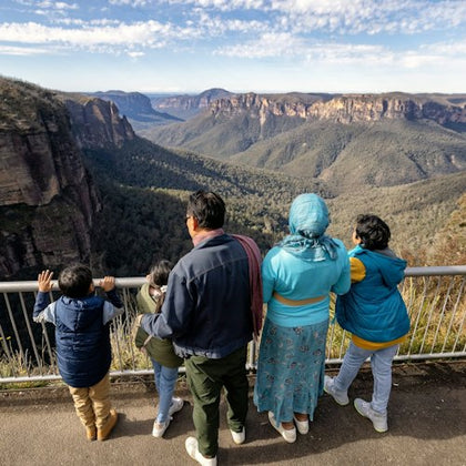 A family of five stands at a railing overlooking a vast canyon landscape with cliffs and forested valleys under a partly cloudy sky.