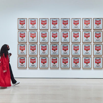 Person in a red coat walks past a wall display of framed Campbell's Soup can artworks arranged in a grid.