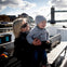 Woman holding a smiling child on a boat with the Tower Bridge in the background on a partly cloudy day.