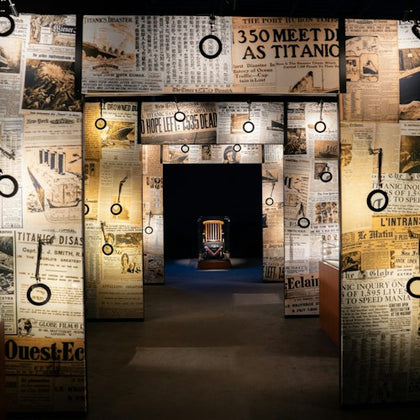 A museum exhibit with walls covered in Titanic newspaper headlines, featuring a spotlighted chair in the background.
