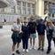 Five people posing together outside a large building, with additional people and filming equipment visible in the background.