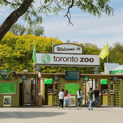Entrance to the Toronto Zoo with signs, ticket booths, and visitors under a clear sky surrounded by greenery.